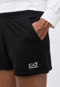 Χέρι που ακουμπά στην τσέπη των μαύρων σορτς EA7 Emporio Armani, εμφανίζοντας το λογότυπο στο μηρό, με φόντο μια λευκή μανσέτα πουκάμισου.