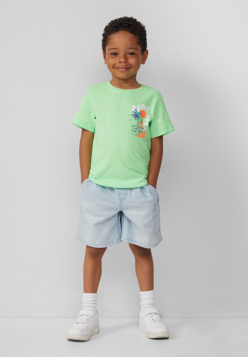 Lichtgroen katoenen t-shirt met korte mouwen en een palmboomafbeelding. Gepaard met lichtblauwe shorts en witte sneakers met zichtbare veters.