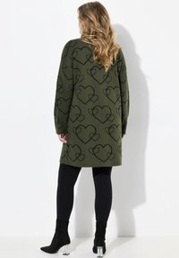 Strickpullovermantel in olivgrün, mit einem schwarzen Herzmuster, langen Ärmeln und einer entspannten Passform, kombiniert mit schwarzen Leggings und Stiefeletten.