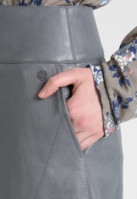 Falda de cuero sintético gris con una cinturilla alta, que presenta un bolsillo lateral y costuras curvadas para un diseño estructurado. Blusa floral visible.