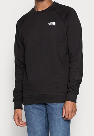 Mann trägt einen schwarzen Langarm-Crewneck-Pullover mit kleinem weißem The North Face-Logo und blauen Jeans.