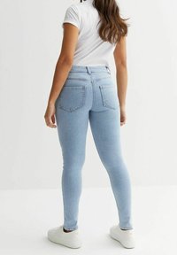 Jeans skinny de color azul claro con cintura alta, dos bolsillos traseros y una textura de mezclilla suave, combinados con zapatillas blancas y una camiseta ajustada.