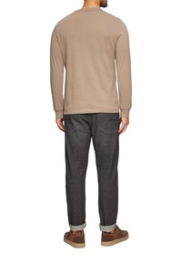 Beige longsleeve shirt met ronde hals, gecombineerd met donkere jeans. Sneakers hebben bruine accenten. De stof lijkt zacht en heeft een ontspannen pasvorm.