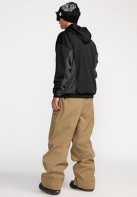 Un sweat à capuche noir avec des accents gris, associé à un pantalon ample beige. Un bonnet noir et des lunettes de ski complètent la tenue.