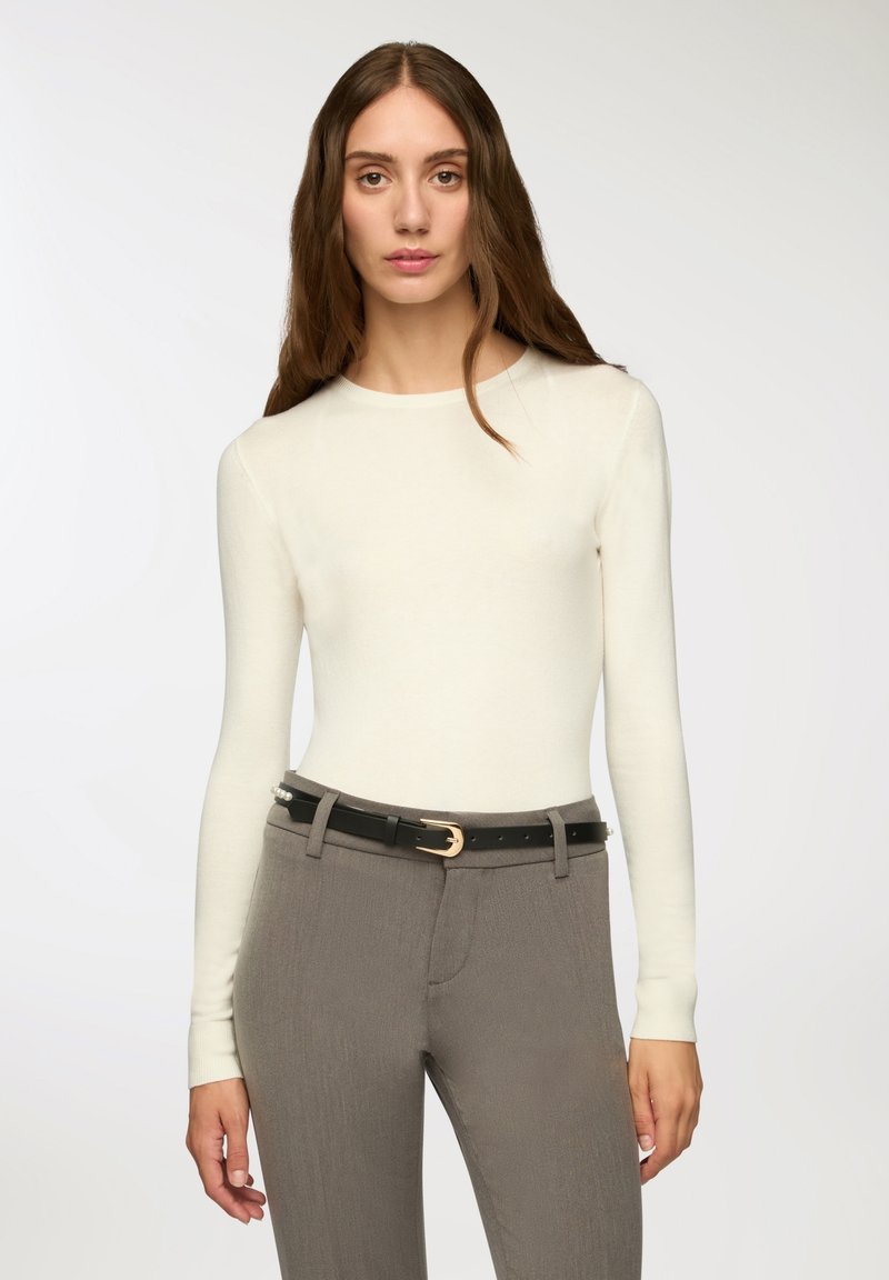 Długi rękaw kremowa bluzka wykonana z miękkiego materiału, zestawiona z dopasowanymi szarymi spodniami. Spodnie wyposażone są w czarny pasek z złotą klamrą.