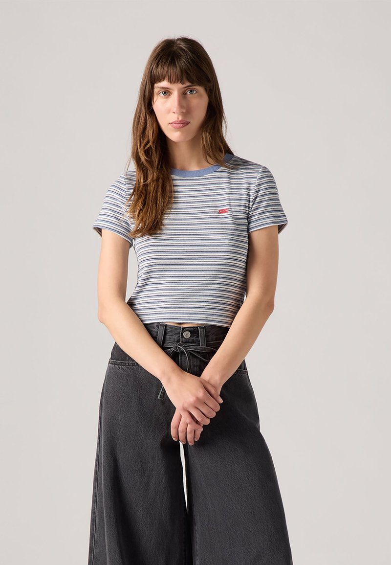 Svītrainā kokvilnas t-kreklā ar zilu apkakli, sarkanais logos uz krūtīm, nēsāta ar augsto jostasvietu melnām platām biksēm, stāvot neitrālā fona.