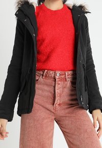 Svart parkajakett med fuskpäls på huven, två sidfickor, dragkedja, bärs över en röd texturerad tröja och ljusrosa jeans.