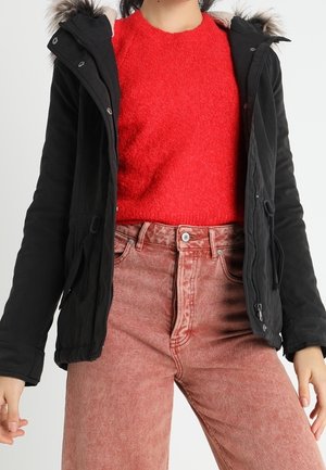Parka nera con cappuccio in pelliccia sintetica, due tasche laterali, chiusura con zip, indossata sopra un maglione rosso a trama e jeans rosa chiaro.