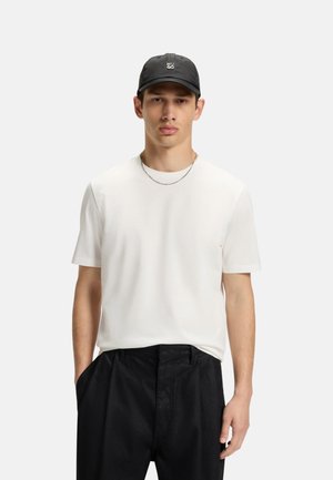 Junger Mann trägt ein weißes kurzärmeliges T-Shirt, schwarze Hose, schwarze Kappe mit Logo und silberne Halskette, steht mit den Händen in den Taschen.