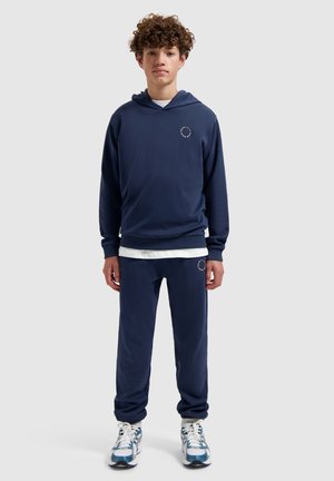 Tienerjongen staand, gekleed in een marineblauwe hoodie en bijpassende broek met witte sneakers, effen lichtgrijze achtergrond.