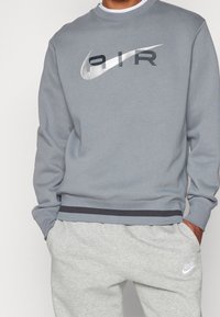 Muž v světle šedých Nike teplácích a šedé mikině s nápisem "AIR" a logem Nike swoosh přes hrudník. Ruce v kapsách.