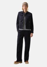 Woman wearing brown patterned headband, clear glasses, navy jacket with pockets, black and white striped shirt, black pants, and brown shoes.