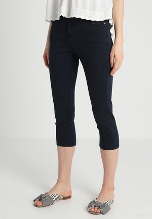 Femme portant un pantalon skinny court bleu foncé, un haut blanc avec un ourlet en dentelle, et des sandales plates rayées à enfiler avec un détail nœud.