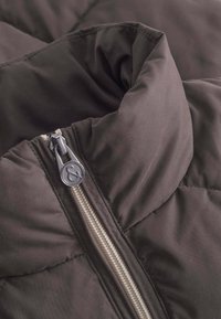 Schwarze gepolsterte Jacke mit hohem Kragen, strukturiertem Stoff und silbernem Reißverschluss mit Logo-Zipper. Nahaufnahme von Kragen- und Reißverschlussdetails.