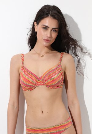 Donna con capelli lunghi e scuri che indossa un bikini colorato con motivo a zigzag in tonalità rosa, arancione e gialla su sfondo bianco.
