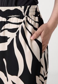 Elasticated waist pants, featuring a black and cream leafy pattern. The fabric is lightweight with a smooth texture. Hand resting in side pocket.