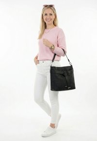 Mujer con suéter rosa, pantalones blancos y zapatillas, llevando un bolso negro de hombro con detalle de hebilla, sonriendo frente a un fondo blanco.