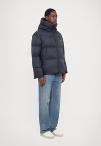 Jachetă puffer de marină, cu secțiuni mari quilted, guler înalt și glugă. Asortată cu blugi bleu deschis și adidași albi.