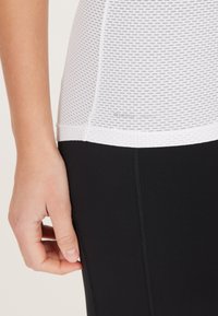 Hvid mesh tanktop med tætsiddende sorte leggings, der har synlige sømme og en glat overflade i overgangen mellem beklædningsstykkerne.