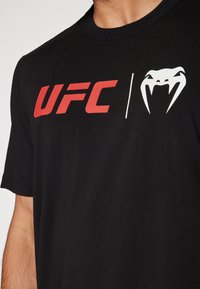 Sort bomulds t-shirt med fed rød "UFC" tekst og et hvidt slangelogo, med rund halsudskæring og korte ærmer.