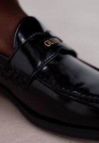Melnas lakota ādas loafers ar noapaļotu pirkstu, ar zelta logotipa akcentu uz siksniņas un redzamām šuvēm.