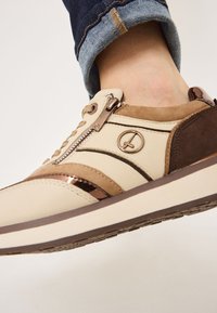 Sneaker beige e marrone con una tomaia texturizzata, dettagli metallici, zip laterale, logo rotondo e strisce a contrasto sui lati.