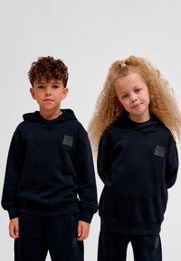 Svarta hoodies med en texturerad lapp, lös passform och ribbade muddar, båda matchande svarta sweatpants. Bakgrunden är enfärgat vit.