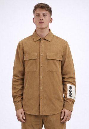 Giovane uomo che indossa una camicia a maniche lunghe color beige in velluto a coste con due tasche sul petto e una toppa bianca con testo nero sul braccio sinistro.