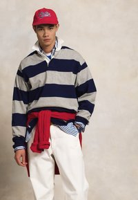 Gray and navy striped long-sleeve polo shirt with a collar, red sweater tied at the waist, white pants, and a red cap with logo.