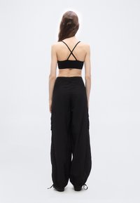 Femme debout, de dos, portant un crop top noir à bretelles croisées et un pantalon cargo noir ample, sur un fond blanc uni.