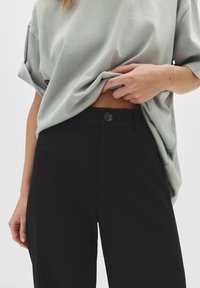 Persoon die een lichtgrijze oversized shirt omhoog heft om zwarte high-waisted broek met knoopsluiting te onthullen.