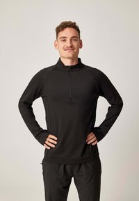 Schwarzer Pullover mit langen Ärmeln, hohem Kragen und Halb-Reißverschluss. Hergestellt aus glattem, elastischem Stoff mit Raglanärmeln für bessere Bewegungsfreiheit.