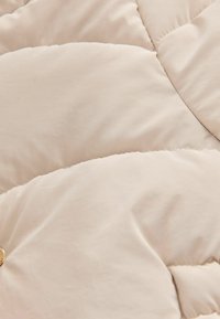 Tissu en duvet de couleur crème avec un design matelassé, présentant une texture lisse et des coutures surpiquées. Un bouton en métal est visible près du bas.