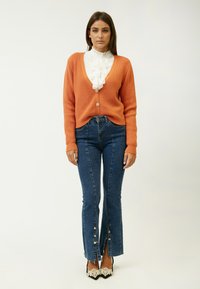 Orange ribbet cardigan med dyb V-udskæring, hvid bluse med flæser under, parret med mørkeblå flare jeans med knapåbninger i kanten.