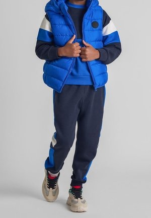 Niebieska pikowana kamizelka z kapturem, granatowa bluza oraz pasujące spodnie dresowe z niebiesko-białymi paskami; całość uzupełniona beżowymi sneakersami.