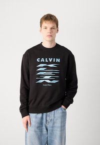 Sweatshirt preto com design gráfico azul em destaque com a palavra "CALVIN" e "Calvin Klein." Apresenta punhos e decote canelados, combinado com jeans azul claro.