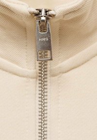 Beige Kapuzenjacke mit durchgehendem Reißverschluss aus silbernem Metall und einem rechteckigen Zipper, auf dem der Markenname "MNG" eingeprägt ist. Texturiertes Material sichtbar.