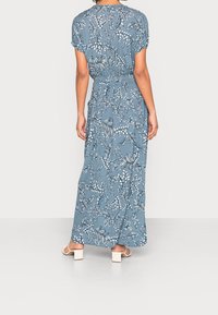 Kvinde iført en blå maxikjole med blomsterprint og korte ærmer samt hvide klodsåler, set bagfra.