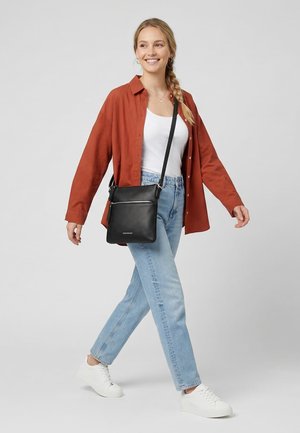 Young woman smiling, walking, wearing rust-colored shirt, white top, blue jeans, white sneakers, and carrying a black crossbody bag.