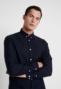Camisa de botones de manga larga en color navy con cuello, botones blancos y bordado sutil del logo en el pecho, hecha de una tela suave.