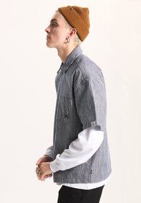 Striped, button-down shirt in navy and white with chest pocket, layered over a gray long-sleeve shirt. Worn with a brown beanie.