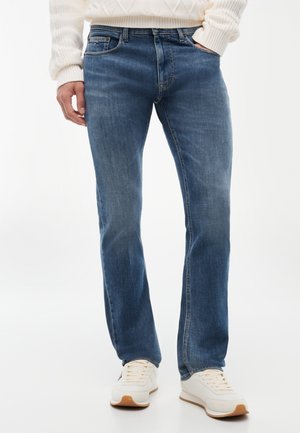 Uomo che indossa jeans dritti a lavaggio medio, maglione bianco di lana e sneakers bianche, in piedi con una mano nella tasca davanti a uno sfondo bianco.