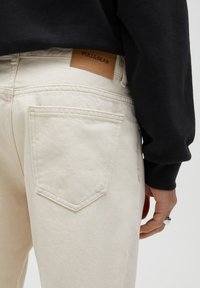 Béžové džíny z denimu s zadní kapsou a hnědou koženou nášivkou s nápisem "PULL&BEAR." Materiál působí měkce a texturovaně.