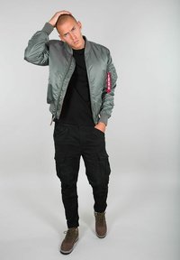 Siva saten bomber jakna s rebrastim manžetama, crna majica, crne cargo hlače i smeđi sportski cipele; model pozira na bijeloj pozadini.