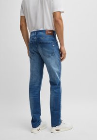 Blå denimjeans med klassisk passform, med en brun logopatch på midjebandet och två bakfickor. Åtföljs av vita sneakers.