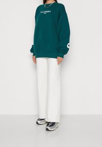 Sweat-shirt surdimensionné bleu sarcelle avec le texte blanc "CALIFORNIA", associé à un pantalon blanc à jambes larges et des chaussures de sport argentées. Manches longues et tissu texturé.