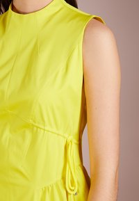 Světlě zelené bezrukávové šaty s vysokým límcem, s vypasovaným pasem, texturovanou tkaninou a bočním detailem na stažení.