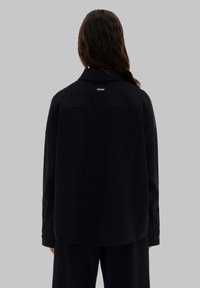 Zwarte shirt met kraag, ontspannen pasvorm, zachte textuur en geknoping manchetten. Heeft een label aan de achterkant van de halslijn.