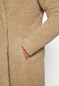 Beige fleecový kabát s texturovaným, nadýchaným povrchem, obsahující boční kapsu a dlouhé rukávy. Materiál vypadá jemně a teple.