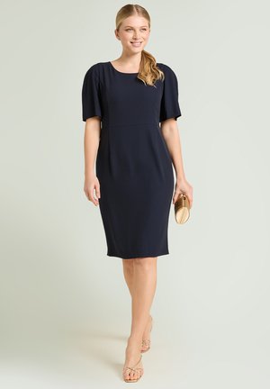 Donna che cammina con sicurezza indossando un vestito blu navy al ginocchio con maniche a sbuffo, tiene in mano una pochette dorata, indossa tacchi dorati con cinturini, sorridendo.